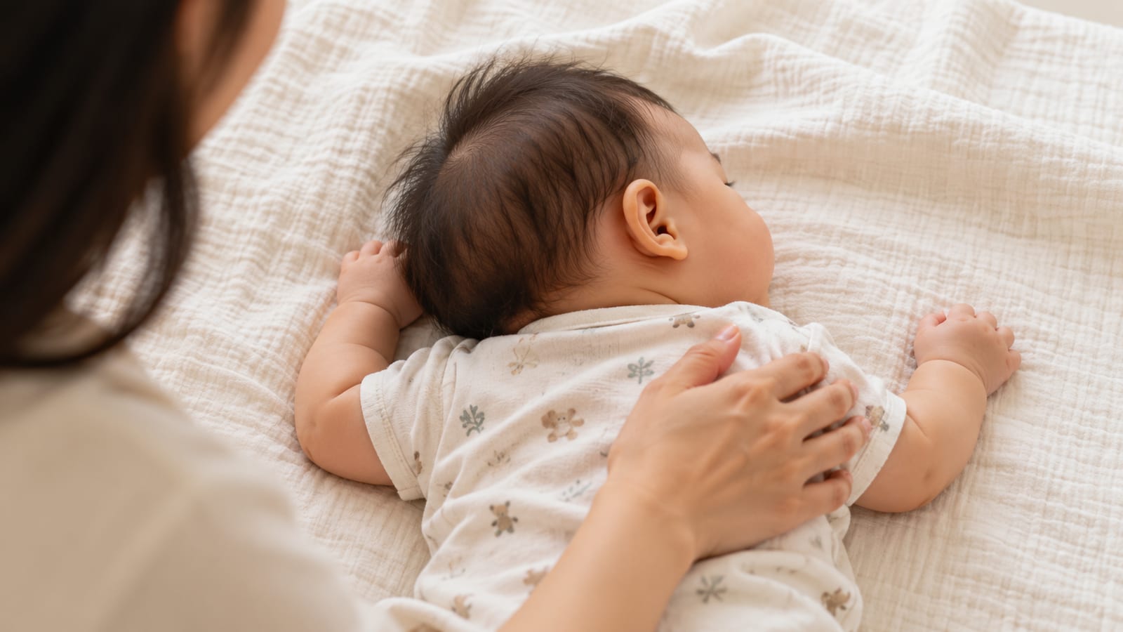 A parent's hand resting lightly on a baby's shoulder as the baby lies with their head to one side