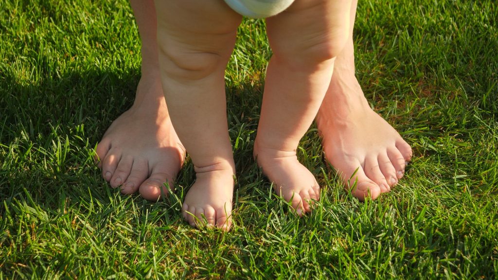A baby standing barefoot on grass with a parent, supporting natural foot and balance development.