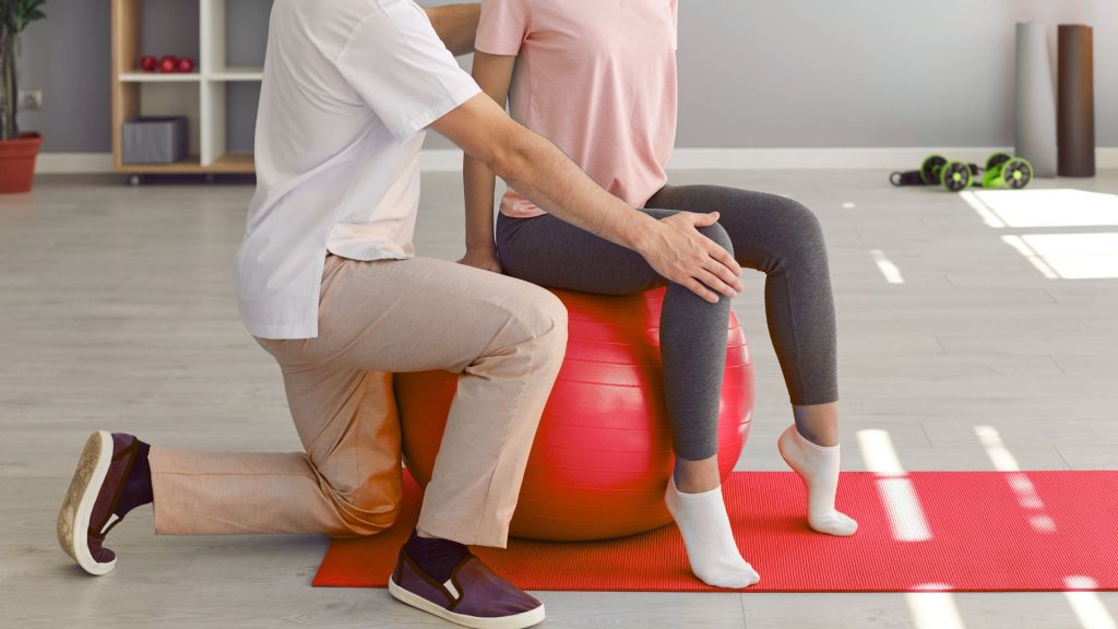 Physiotherapist supporting a woman seated on a red stability ball during guided pelvic floor training for bladder control.