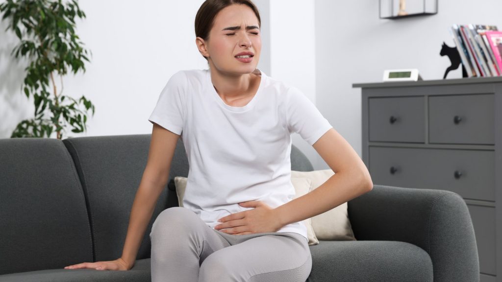 Woman sitting on a sofa holding her lower abdomen in pain, representing symptoms of urinary incontinence.
