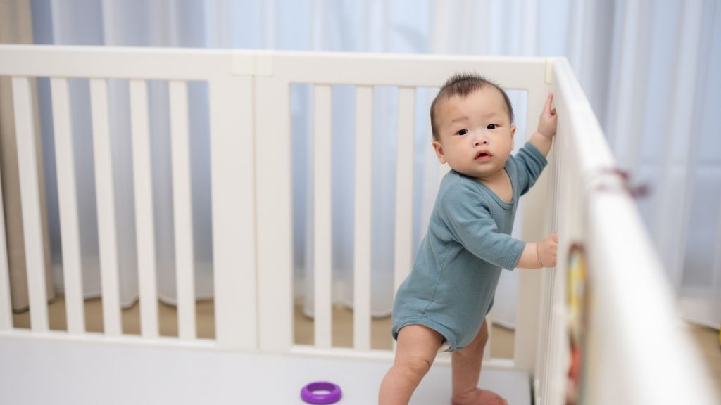 A baby holding onto a playpen rail while standing and practising early cruising, an important pre-walking milestone.