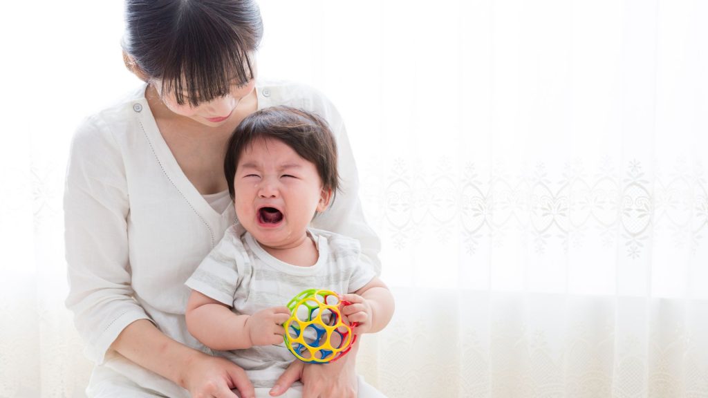 A crying baby sitting on a parent’s lap holding a toy ball, possibly indicating sensory overwhelm or difficulty engaging in play.