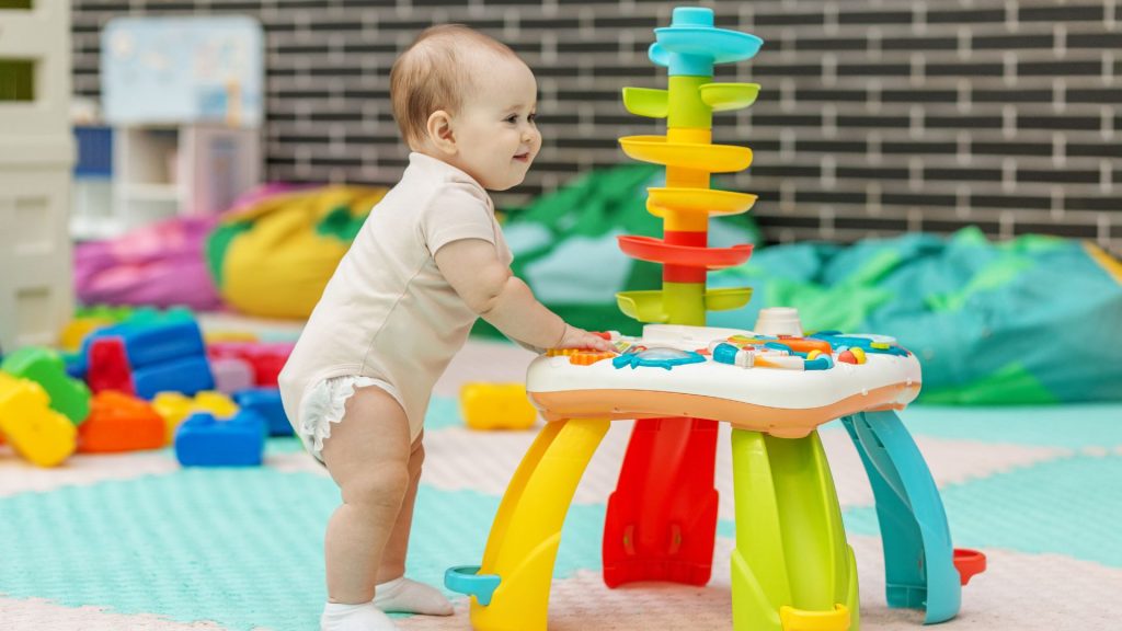 A baby standing and holding onto an activity table while playing, developing balance and early cruising skills.