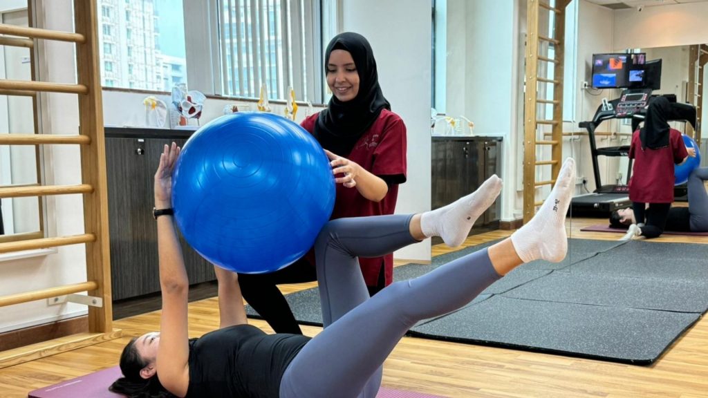 Physiotherapist guiding a woman through a core stability exercise using a fitness ball, part of a pelvic floor physiotherapy program designed to support urine incontinence treatment in Singapore.
