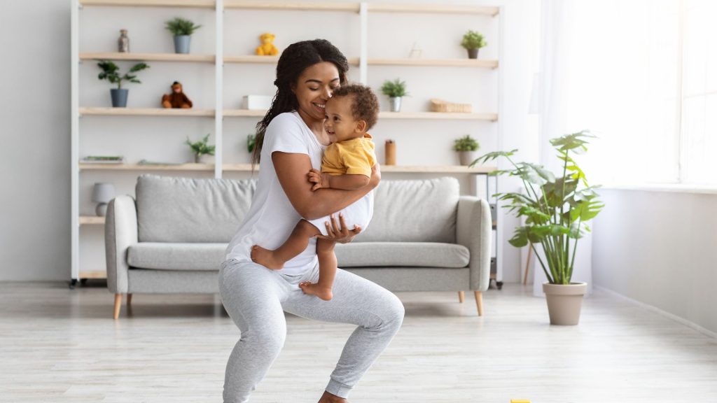 Mum doing squats at home while carrying toddler, smiling and enjoying postnatal movement