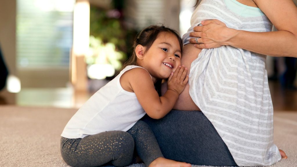 Young girl smiling while listening to her pregnant mum’s belly with love and curiosity