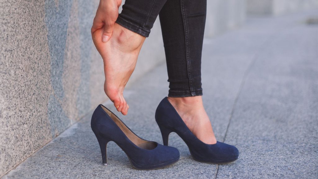 Woman removing high heels while rubbing her sore foot on a concrete walkway.