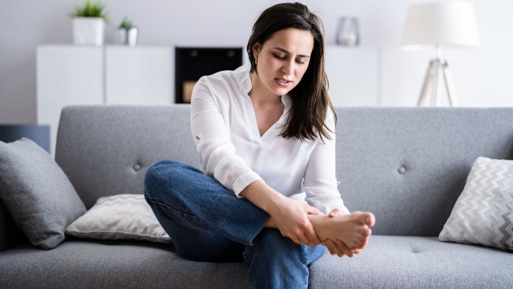Woman sitting on a sofa massaging her painful foot, showing discomfort from prolonged standing or tight shoes.