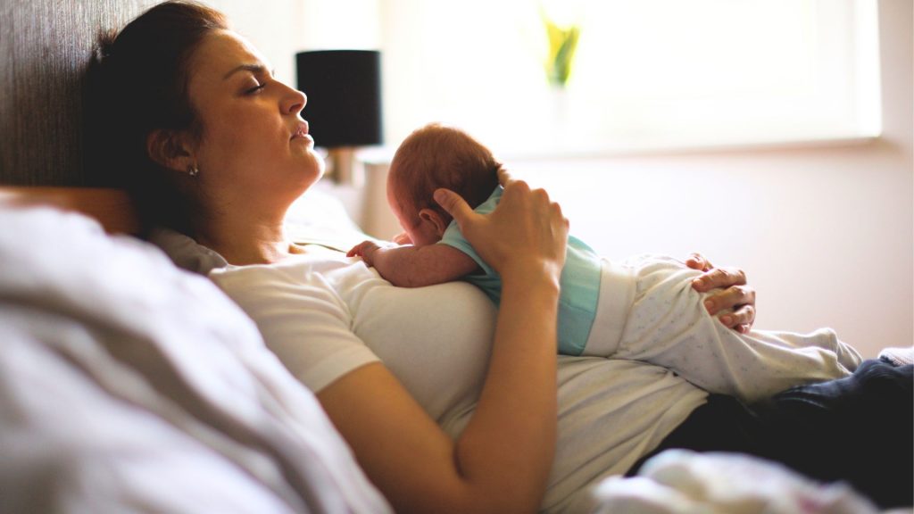 Tired postpartum mother lying in bed with eyes closed, holding a newborn baby on her chest in a quiet, softly lit room