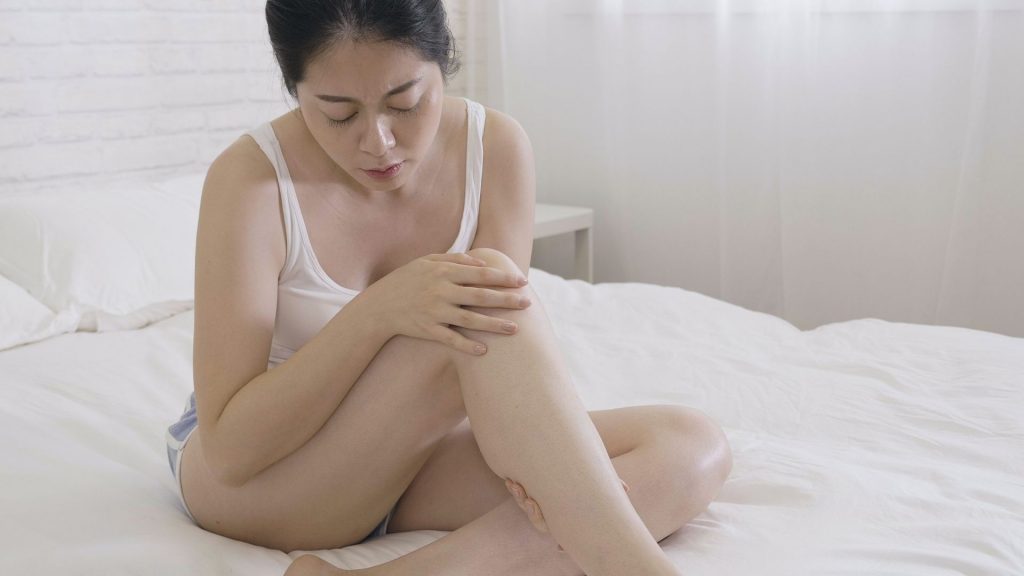 Asian woman sitting on a bed holding her calf in discomfort, showing common morning foot and leg pain in women.