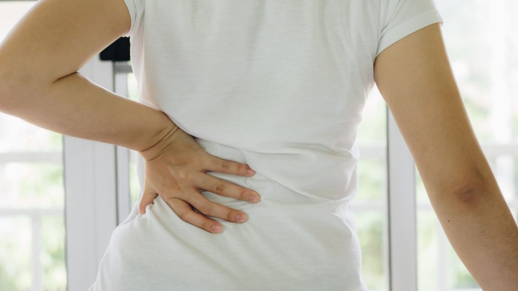 Person wearing a white shirt holding their lower back with one hand, showing signs of discomfort or pain, possibly from posture strain or postpartum recovery