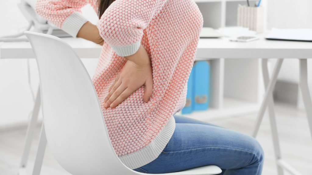 Woman in a pink sweater sitting at a desk with hands on her lower back, showing signs of back pain from prolonged sitting