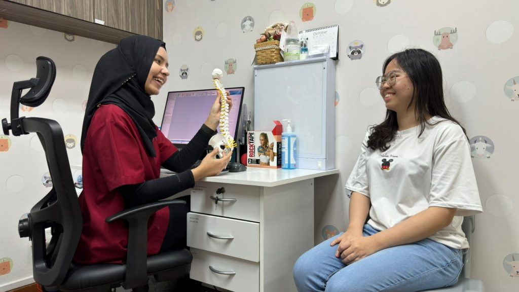 Smiling female physiotherapist in a red uniform explaining spinal anatomy to a young woman during a consultation in a child-friendly clinic room