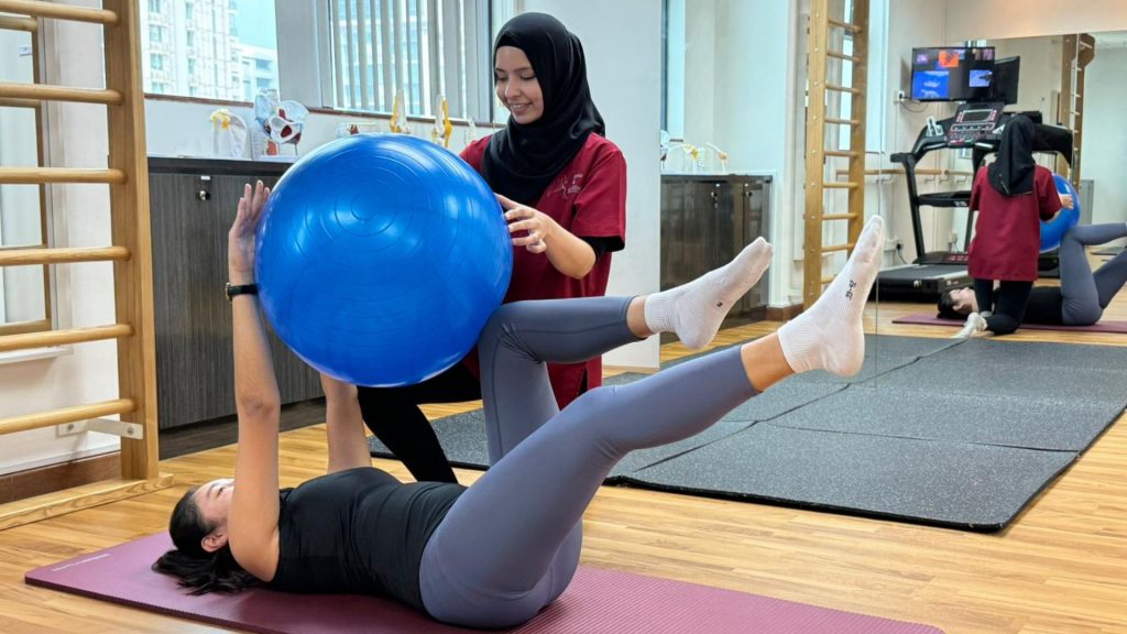 Physiotherapist guiding a woman through a clinical Pilates core exercise using a blue stability ball during a supervised session in a rehabilitation gym