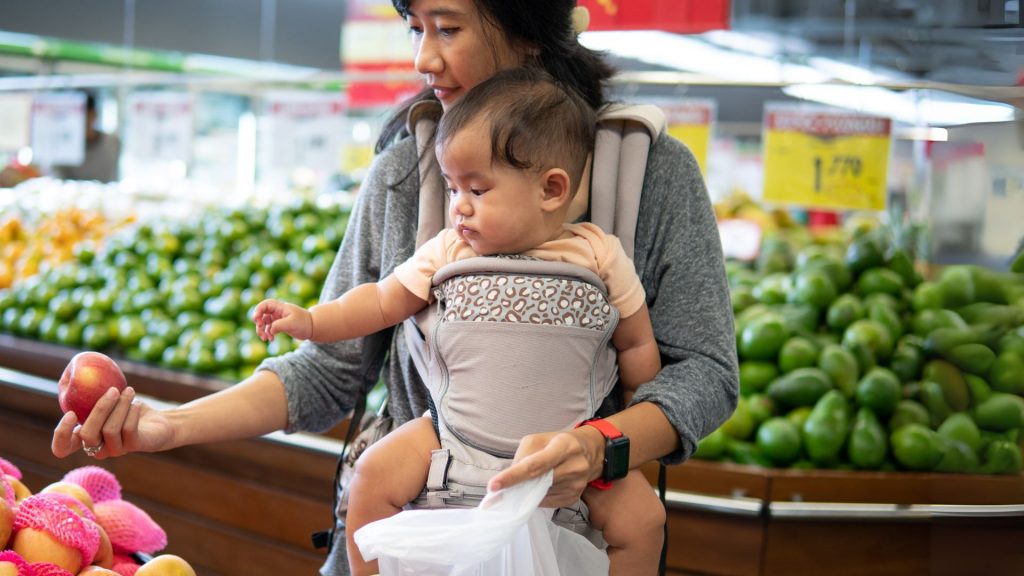 Mother carrying baby in a front-facing carrier while selecting apples at a grocery store produce section