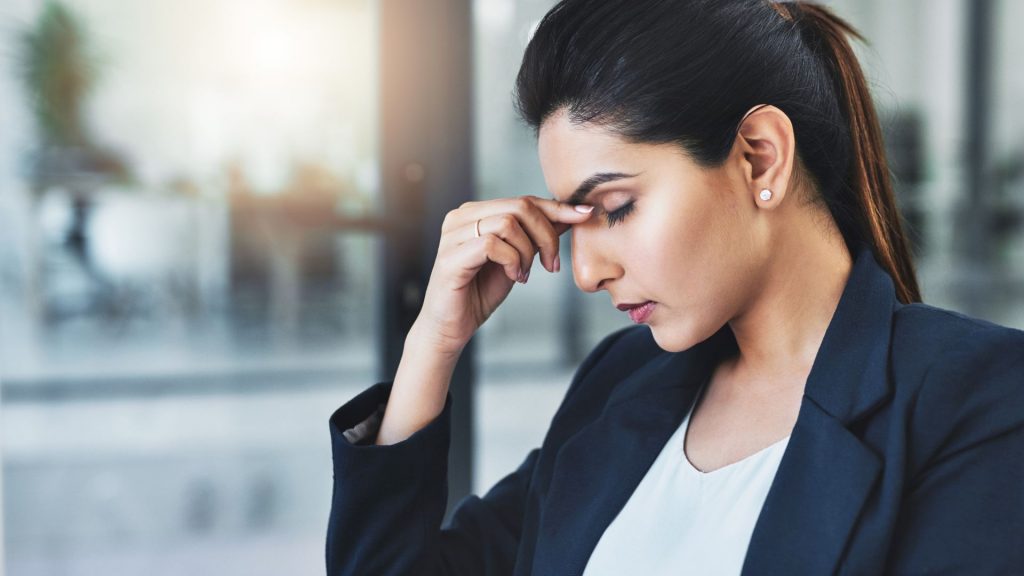 Young woman in office attire holding her forehead in stress, reflecting the emotional toll of urinary incontinence.