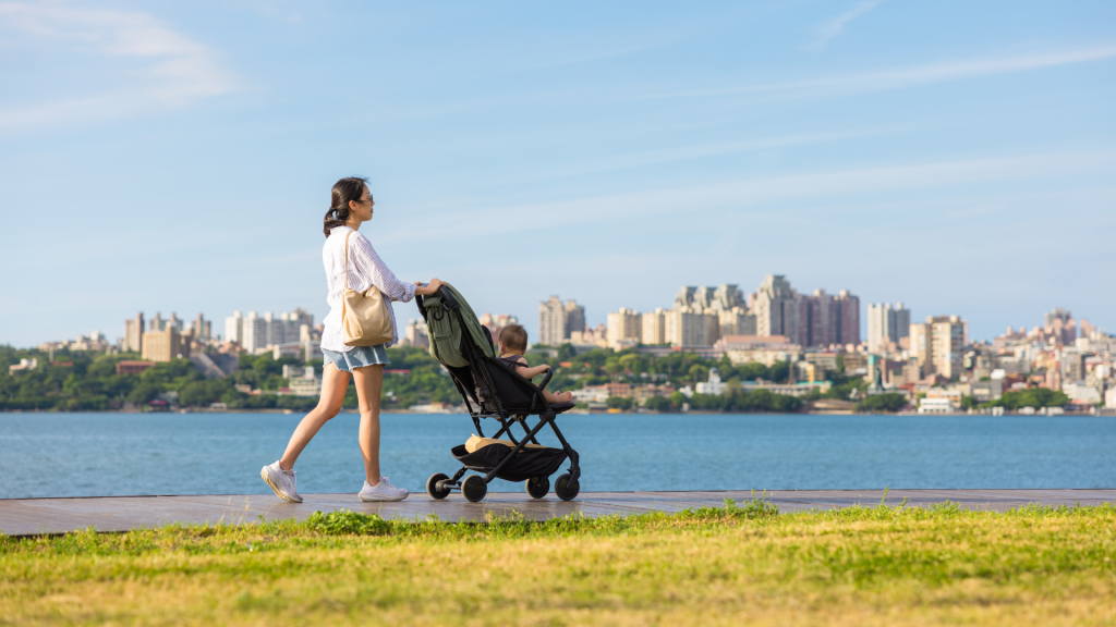 Mother walking along a waterfront while pushing a baby stroller, representing gentle postpartum activity that can affect pelvic girdle and lower back comfort.